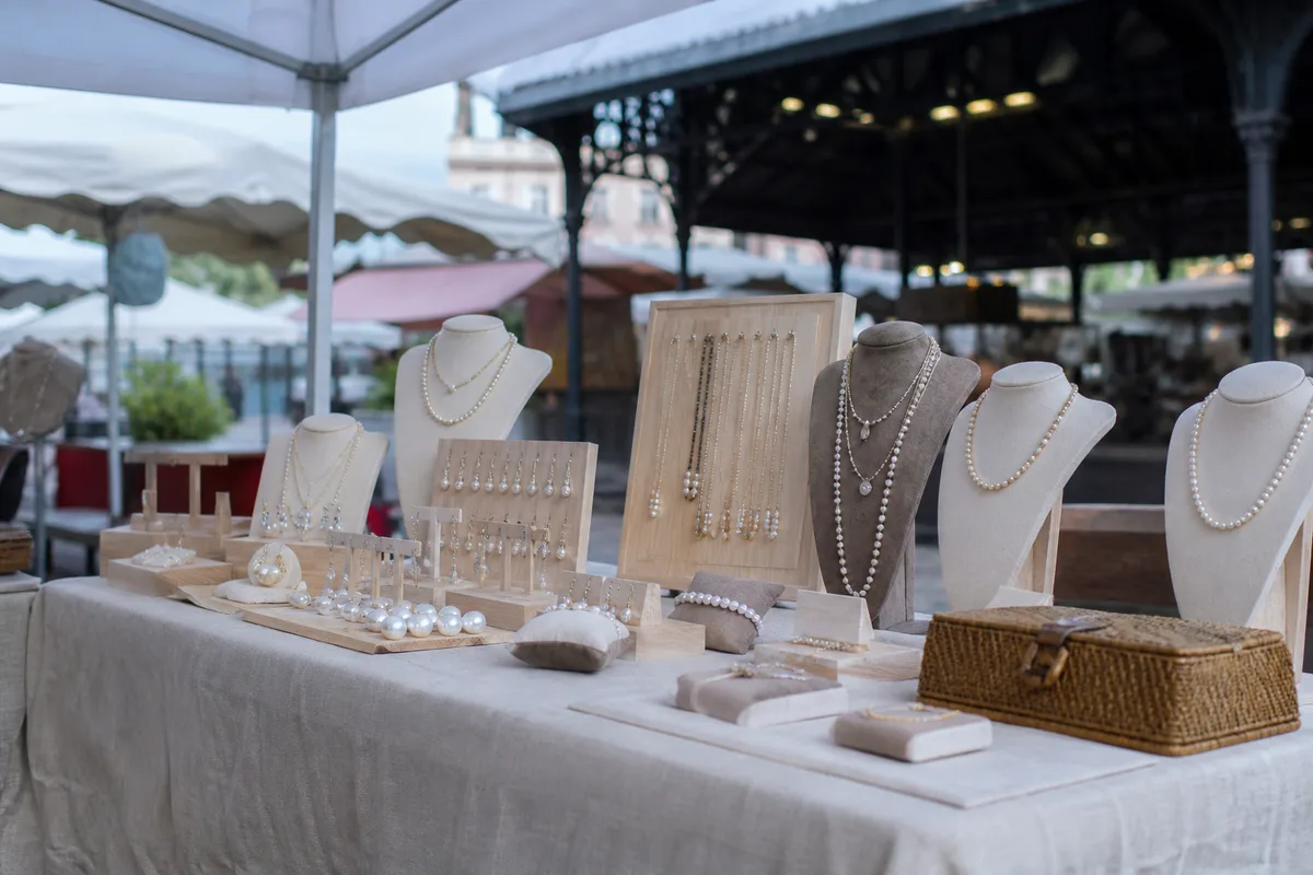 Stand de marché de créateurs au petit matin, présentoirs en bois clair garnis de colliers de perles, mains posant une étiquette manuscrite